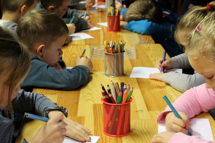 Enfants assis autour d'une grande table d'école et qui dessinent avec des crayons de couleur